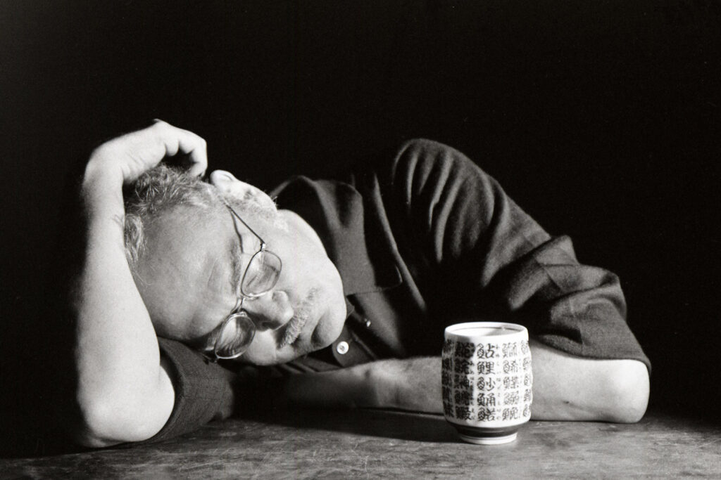 Eugene Smith at a table, lays his head down on his arm, eyes closed. A teacup with kanji characters site in front of him.